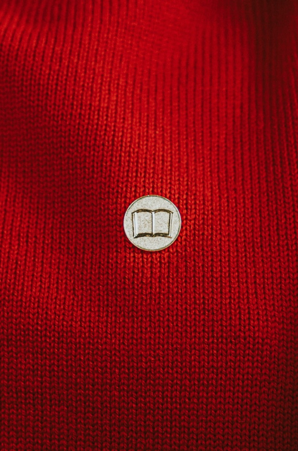Image of a round goldtone pin with a relief image of a an open book pinned to a red sweater.